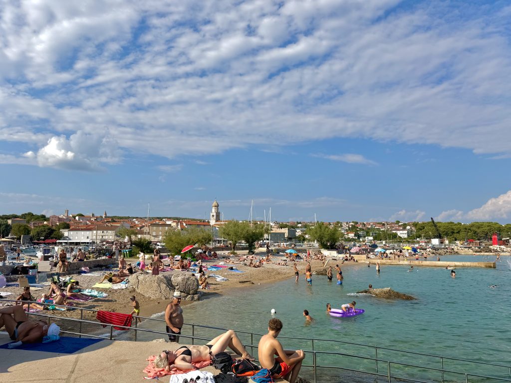Belebter Kieselstrand Porporela in Krk Stadt mit Badegästen, türkisblauem Wasser und Blick auf die Altstadt