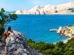 Eine Wanderin mit Rucksack steht auf einem Felsvorsprung und blickt über die raue Küstenlandschaft der Insel Krk auf das blaue Adriatische Meer.