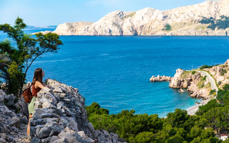 Eine Wanderin mit Rucksack steht auf einem Felsvorsprung und blickt über die raue Küstenlandschaft der Insel Krk auf das blaue Adriatische Meer.