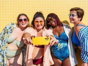 Vier lachende junge Frauen im sommerlichen Outfit machen gemeinsam ein Selfie mit einem gelben Smartphone vor einer sonnigen Wand.