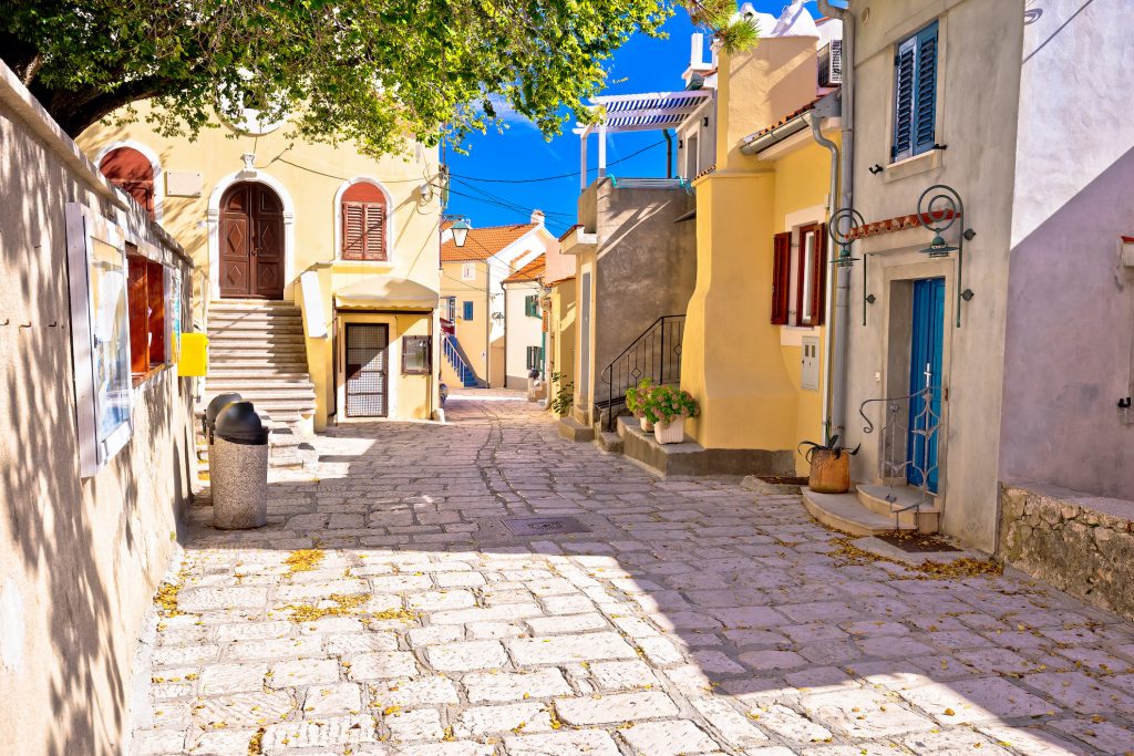 Sonnige und malerische Gasse in der Altstadt von Baška auf der Insel Krk mit farbenfrohen Häusern. Solche beliebten Ferienunterkünfte benötigen ab 2026 zwingend die neue kroatische Registrierungsnummer, um auf Buchungsportalen zu bleiben.