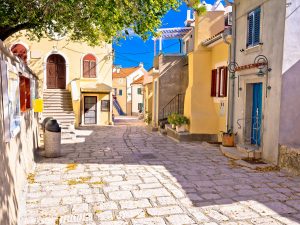 Sonnige und malerische Gasse in der Altstadt von Baška auf der Insel Krk mit farbenfrohen Häusern. Solche beliebten Ferienunterkünfte benötigen ab 2026 zwingend die neue kroatische Registrierungsnummer, um auf Buchungsportalen zu bleiben.