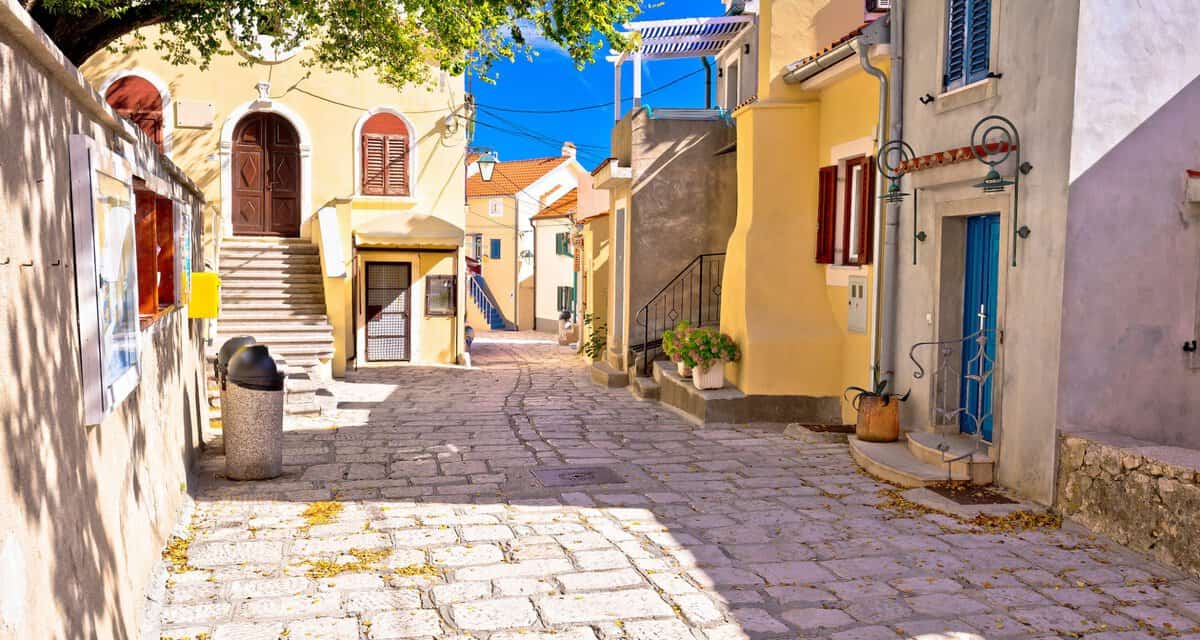 Sonnige und malerische Gasse in der Altstadt von Baška auf der Insel Krk mit farbenfrohen Häusern. Solche beliebten Ferienunterkünfte benötigen ab 2026 zwingend die neue kroatische Registrierungsnummer, um auf Buchungsportalen zu bleiben.