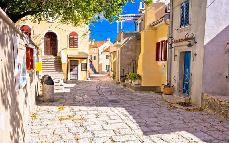 Sonnige und malerische Gasse in der Altstadt von Baška auf der Insel Krk mit farbenfrohen Häusern. Solche beliebten Ferienunterkünfte benötigen ab 2026 zwingend die neue kroatische Registrierungsnummer, um auf Buchungsportalen zu bleiben.