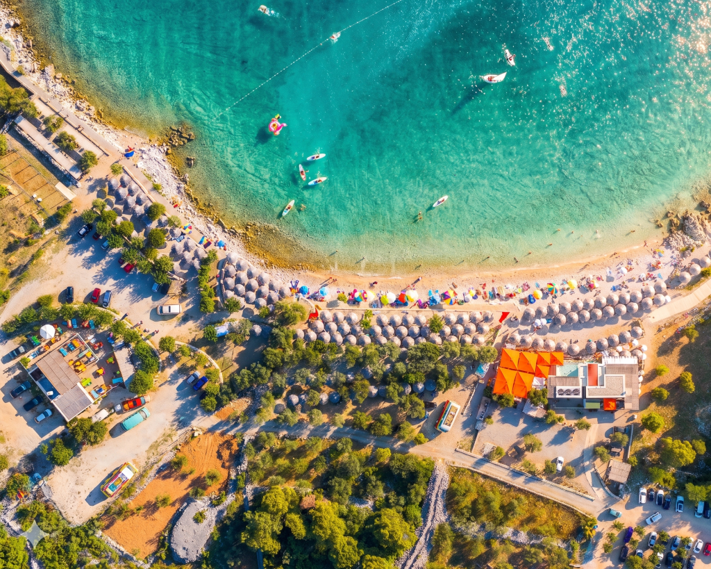 Luftaufnahme vom Medane Beach in Punat auf der Insel Krk mit der türkisfarbenen Bucht, der grünen Liegewiese unter Olivenbäumen und der Beach Bar.