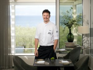 Chefkoch Marin Pleše steht lächelnd in seinem Restaurant Vila Rova. Er trägt eine weiße Kochjacke, auf der sein Name und das JRE-Logo eingestickt sind. Er stützt sich leicht an einem elegant gedeckten Tisch ab, während durch die großen Fenster im Hintergrund das blaue Meer der Kvarner-Bucht zu sehen ist.