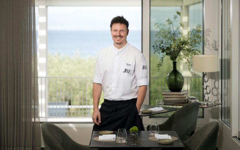 Chefkoch Marin Pleše steht lächelnd in seinem Restaurant Vila Rova. Er trägt eine weiße Kochjacke, auf der sein Name und das JRE-Logo eingestickt sind. Er stützt sich leicht an einem elegant gedeckten Tisch ab, während durch die großen Fenster im Hintergrund das blaue Meer der Kvarner-Bucht zu sehen ist.